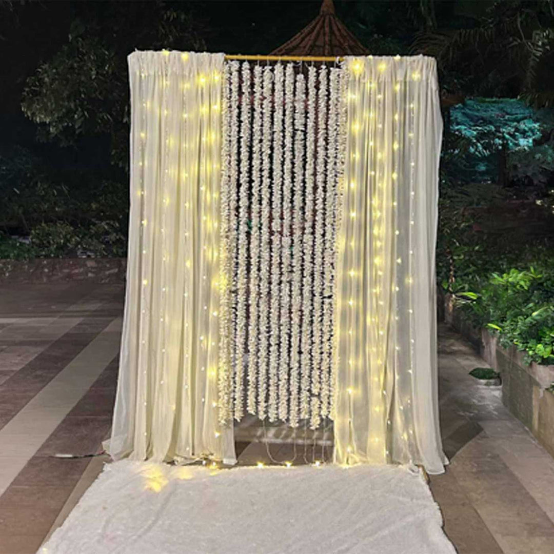 White Floral Garland Backdrop Decor Kit With Off-White Crepe Curtains & Fairy Lights For Weddings