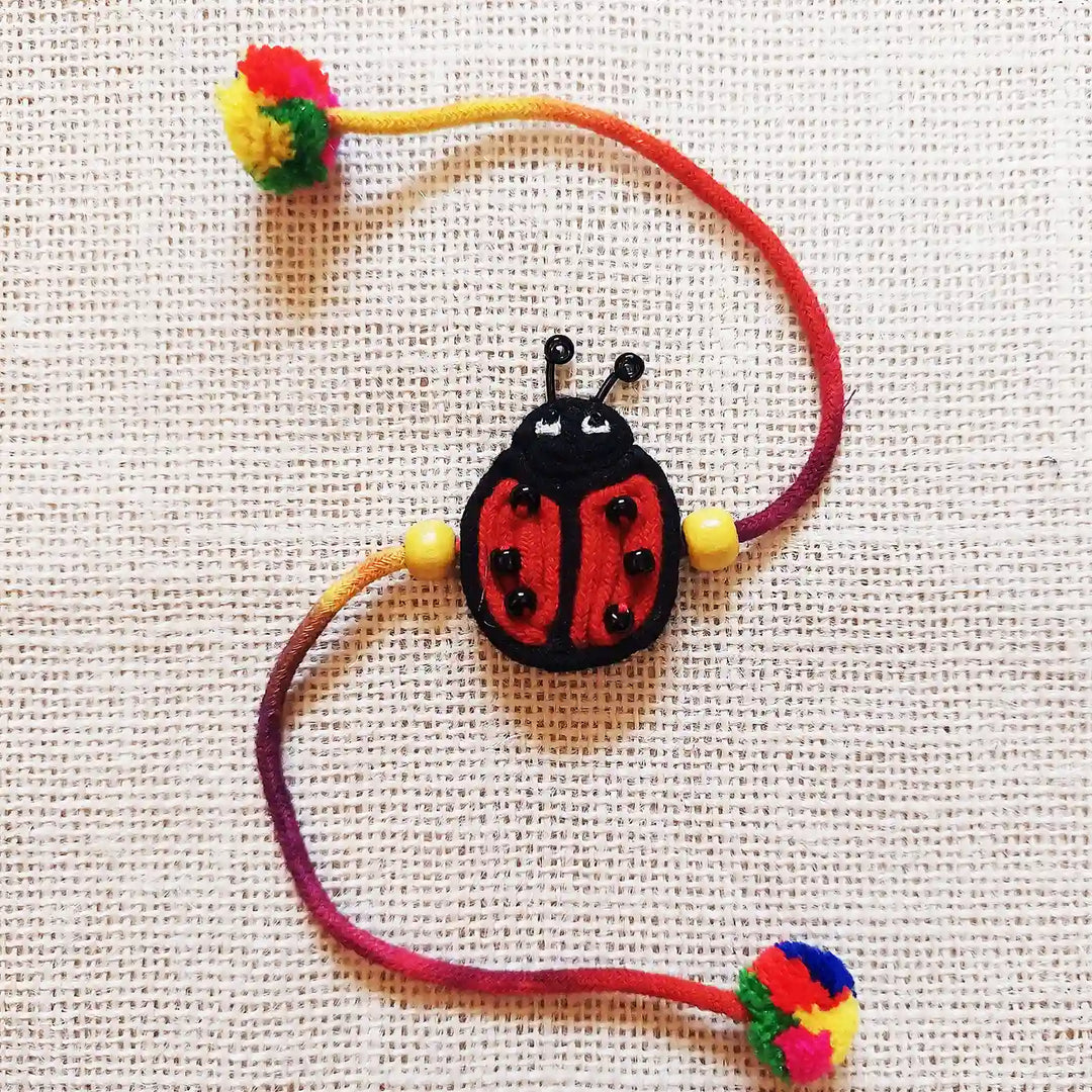 Handmade Dori Work Ladybird Kids Rakhi With Roli Chawal - Front View with Fabric Background