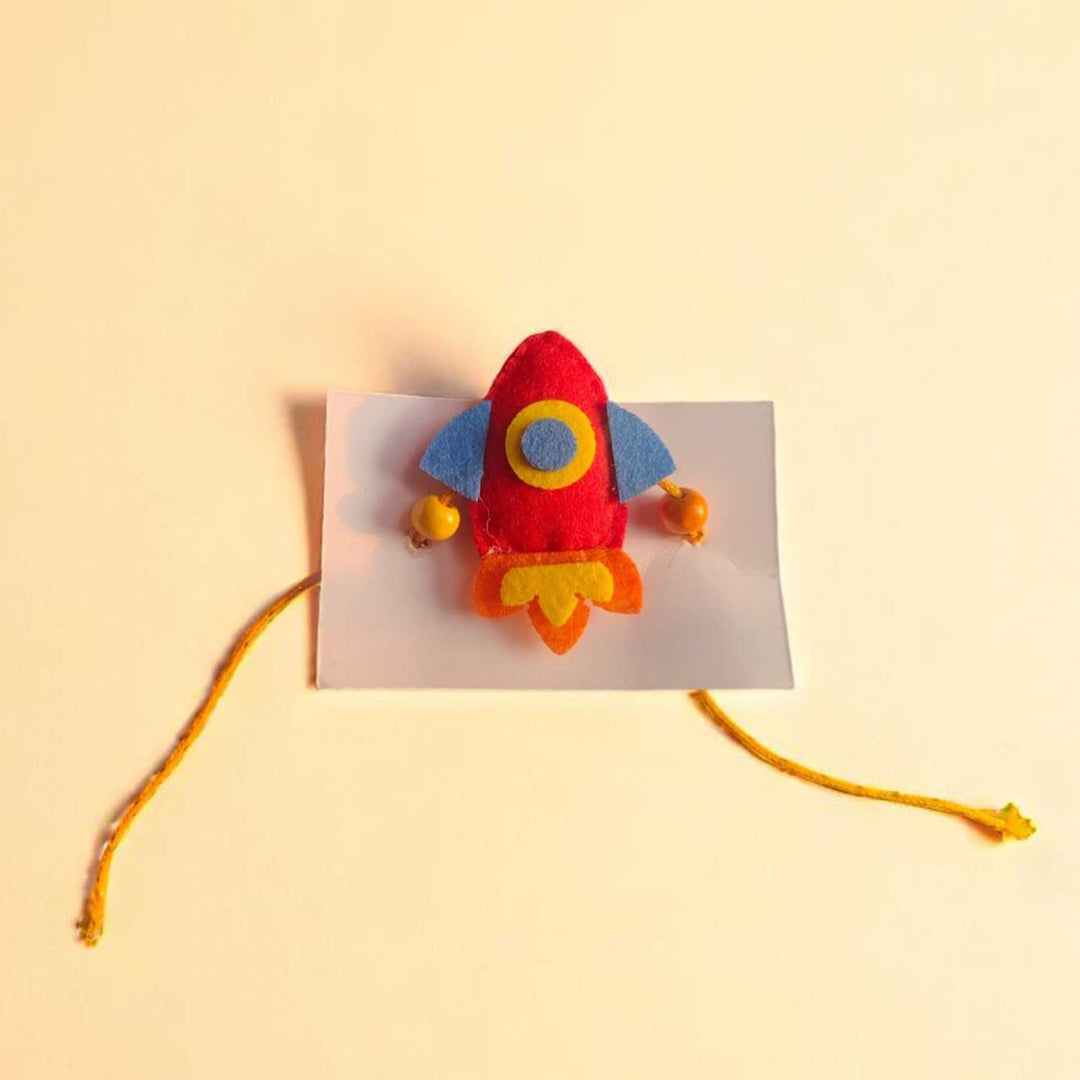 Handmade Beaded Rocket Theme Felt Rakhi With Roli Chawal For Kids - Top View on Cardboard