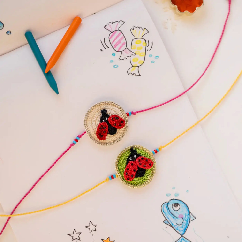 Handmade Crochet Beetle Theme Kids Rakhi With Roli Chawal Set Of 2 - Overhead view with colorful drawing background