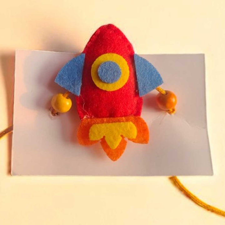 Handmade Beaded Rocket Theme Felt Rakhi With Roli Chawal For Kids - Close-up Front View on Cardboard