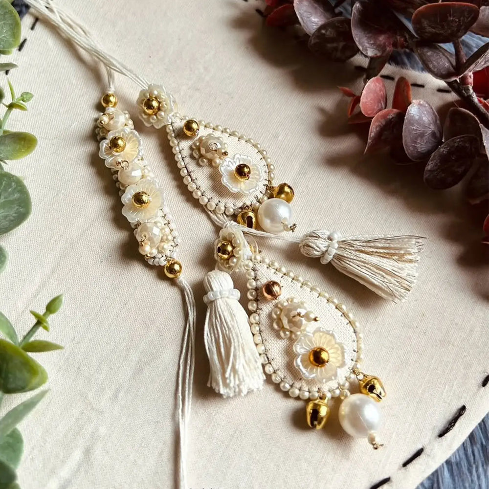 Hand Embroidered Beaded Ivory Bhaiya Bhabhi Rakhi With Roli Chawal Set Of 2 - Close-up of Earrings on Fabric