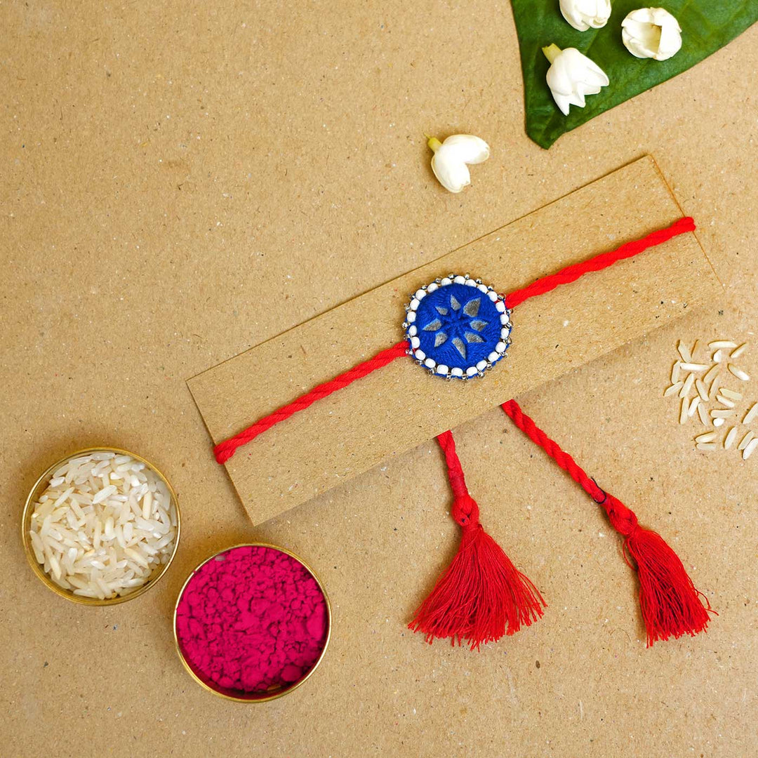 Handmade Mirror Work Kajal Blue Red Rakhi With Roli Chawal - Flat lay with rice and color powder