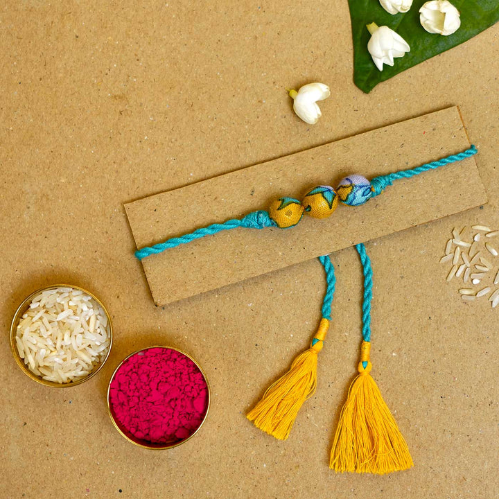 Handmade Turquoise Upcycled Fabric Rakhi With Roli Chawal - Thread Displayed on Cardboard
