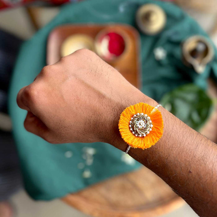 Handmade Zardozi Surajmukhi Rakhi With Wooden Box And Roli Chawal Set Of 2 - Close-up on wrist orange
