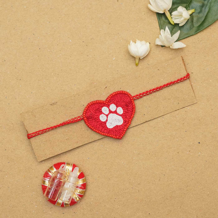 Handmade Beaded Paw Heart Rakhi With Roli Chawal - On-card packaging with flowers and diya