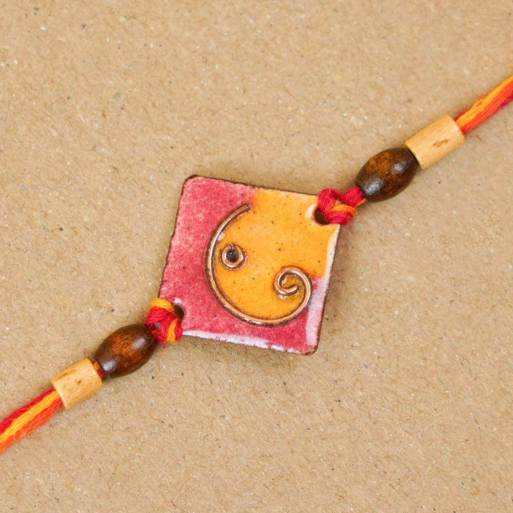 Handmade Square Ganesh Copper Enamel Rakhi With Chocolates And Roli Chawal - Close-up of orange rakhi detail