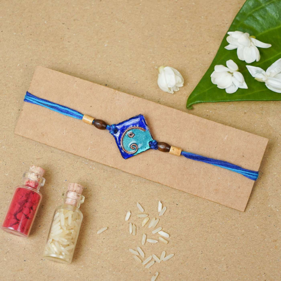 Handmade Square Ganesh Copper Enamel Rakhi With Chocolates And Roli Chawal - Blue rakhi on cardboard packaging