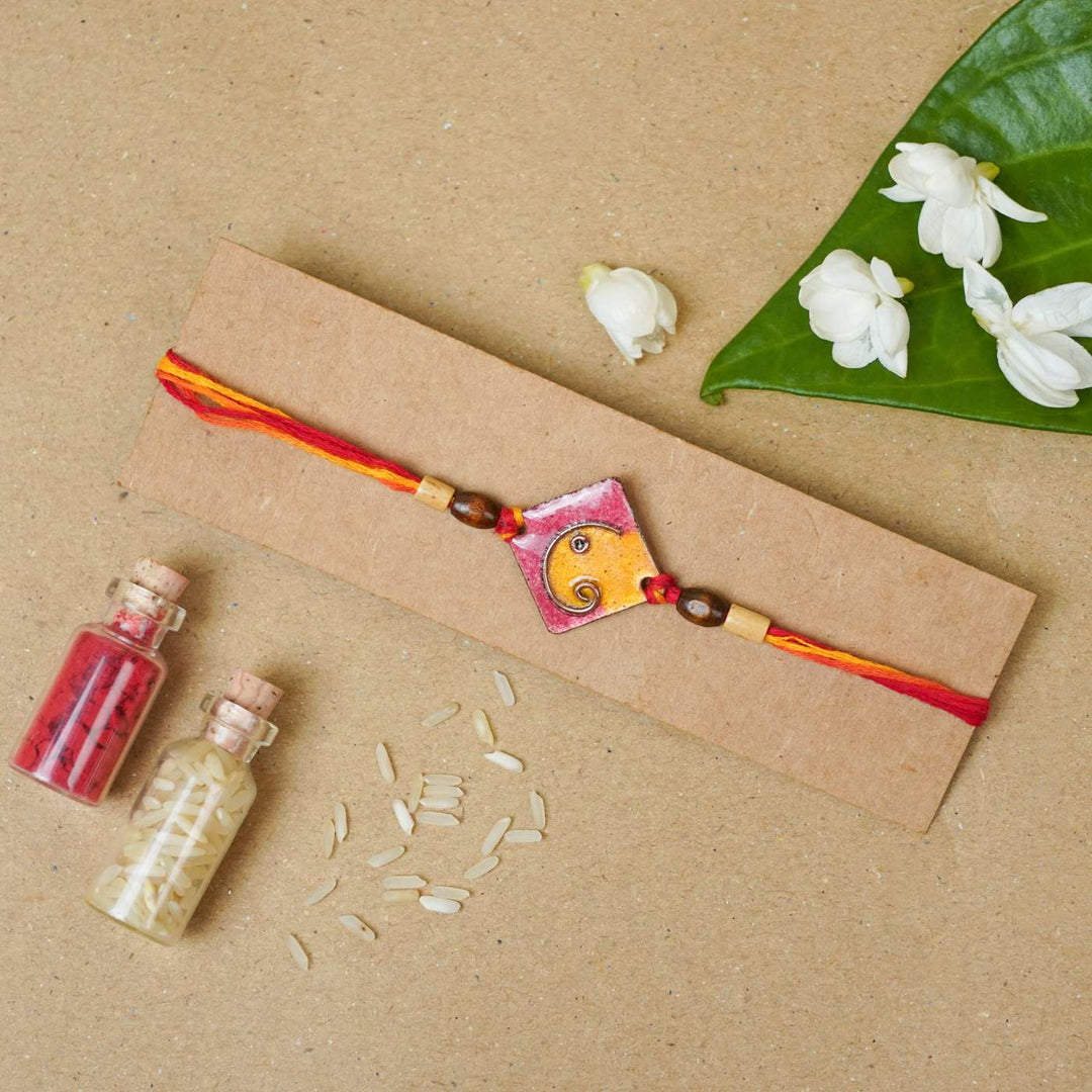 Handmade Square Ganesh Copper Enamel Rakhi With Chocolates And Roli Chawal - Orange rakhi on cardboard packaging