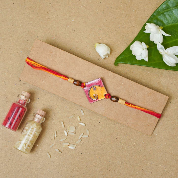 Handmade Square Ganesh Copper Enamel Rakhi With Chocolates And Roli Chawal - Orange rakhi on cardboard packaging