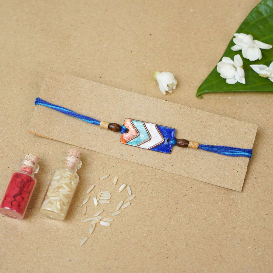 Handmade Arrows Copper Enamel Rakhi With Chocolates And Roli Chawal - Flat Lay on Cardboard Display