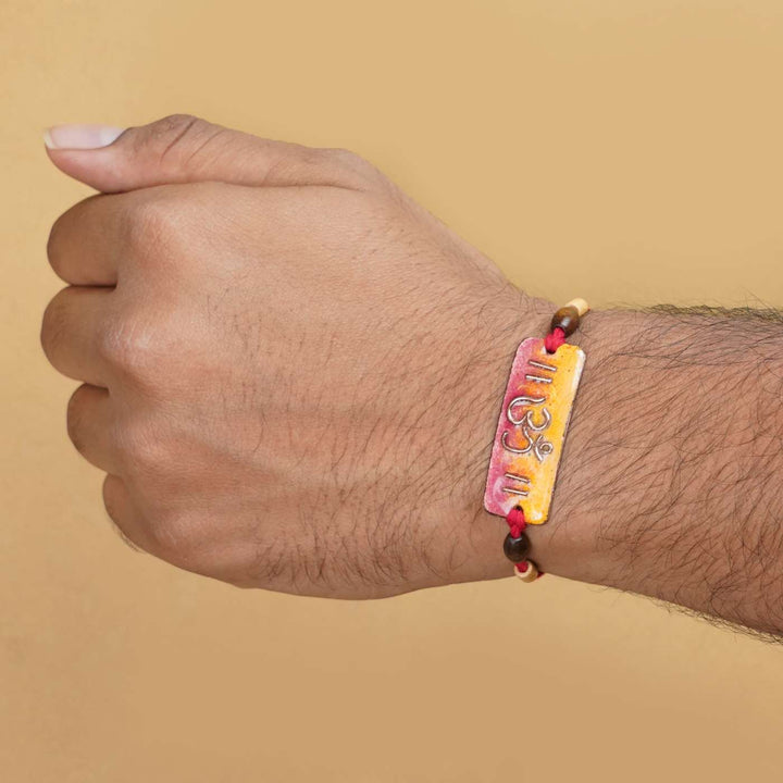 Handmade Rectangle Om Copper Enamel Rakhi With Chocolates And Roli Chawal - On Wrist Model Display