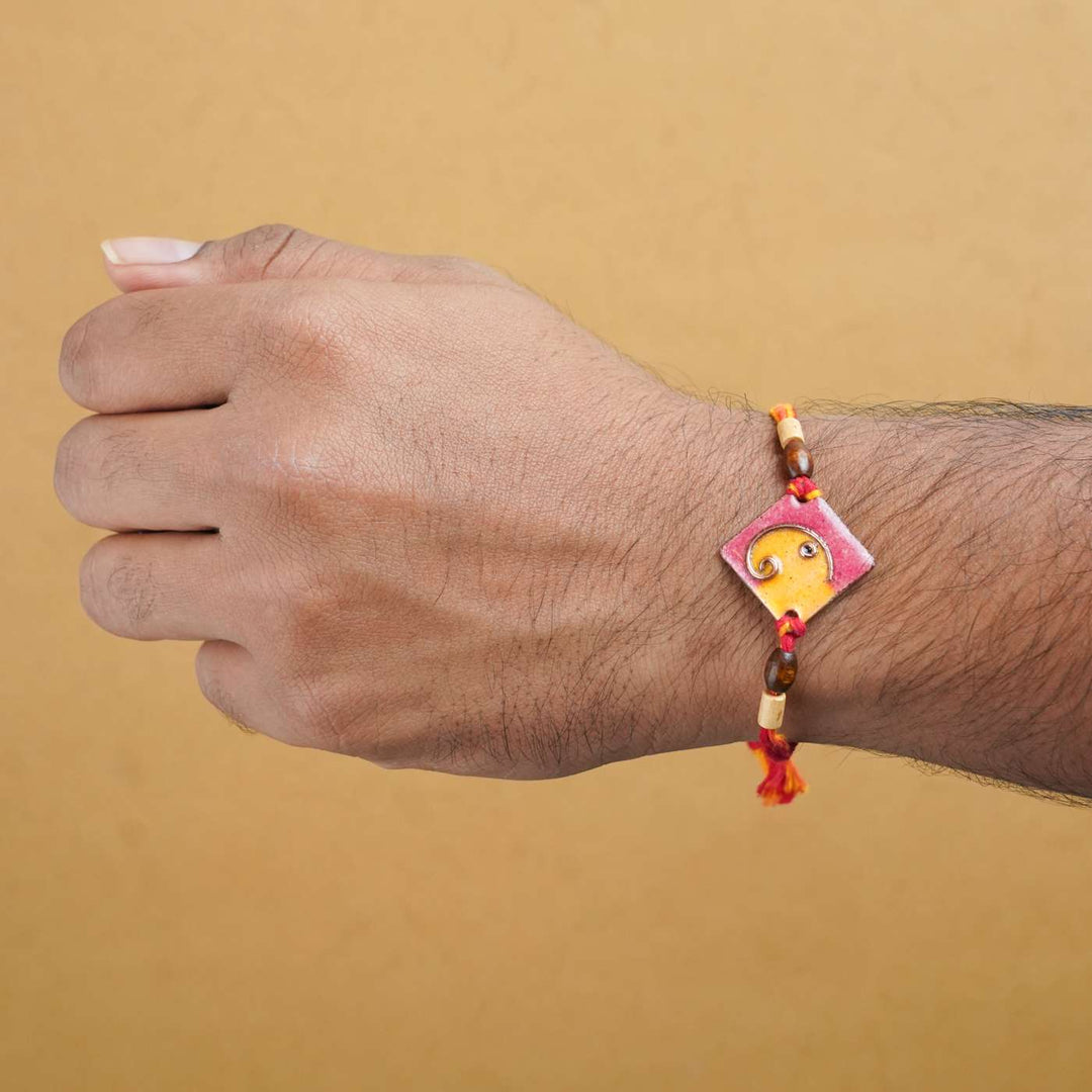 Handmade Square Ganesh Copper Enamel Rakhi With Chocolates And Roli Chawal - Orange rakhi worn on wrist