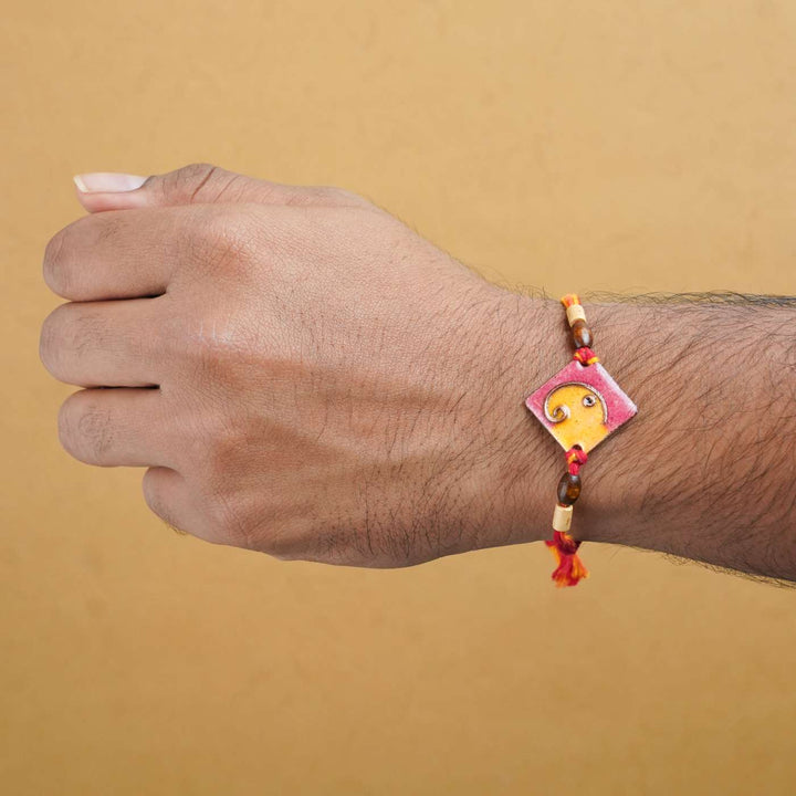 Handmade Square Ganesh Copper Enamel Rakhi With Chocolates And Roli Chawal - Orange rakhi worn on wrist