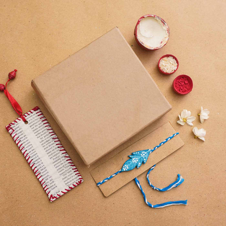 Handmade Ecofriendly Blue Feather Recycled Paper Rakhi With Roli Chawal - Flat lay with packing elements