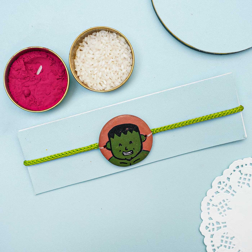 Hand Painted Hulk Terracotta Rakhi With Roli Chawal For Kids - Styled Product Flat Lay with Accessories